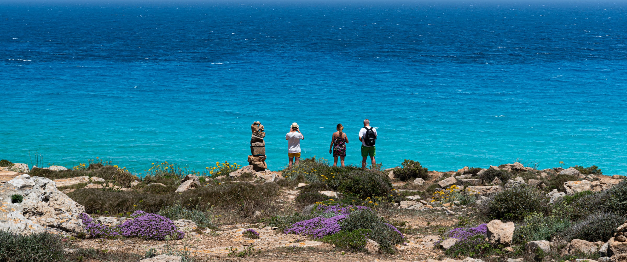 Four travellers standing on a cape above a turquoise sea, wildflowers at their feet (photo: Takahiro Mitsui)