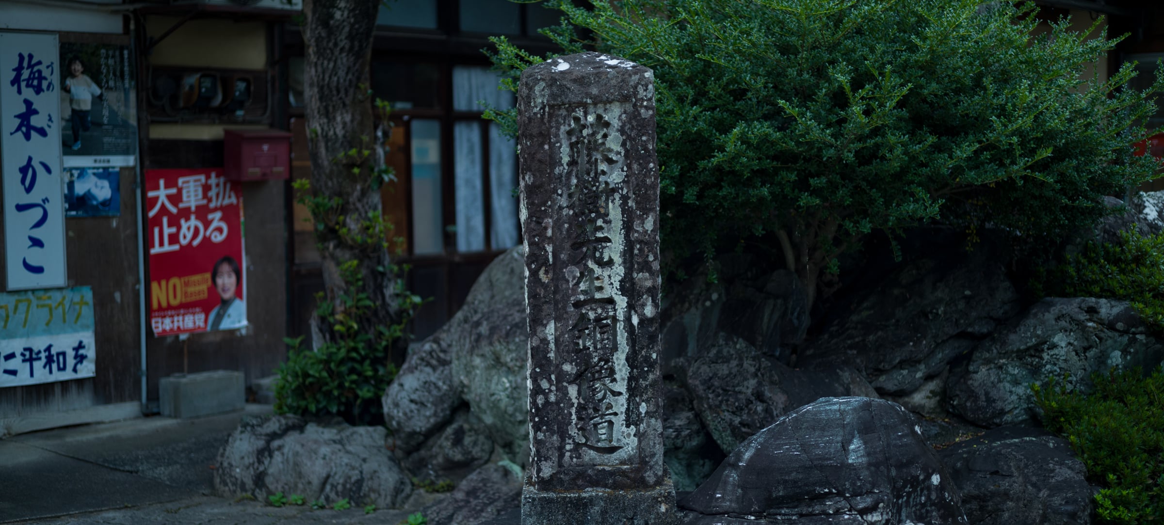 An old stone marker standing beside rocks at a street corner (photo: Takahiro Mitsui)