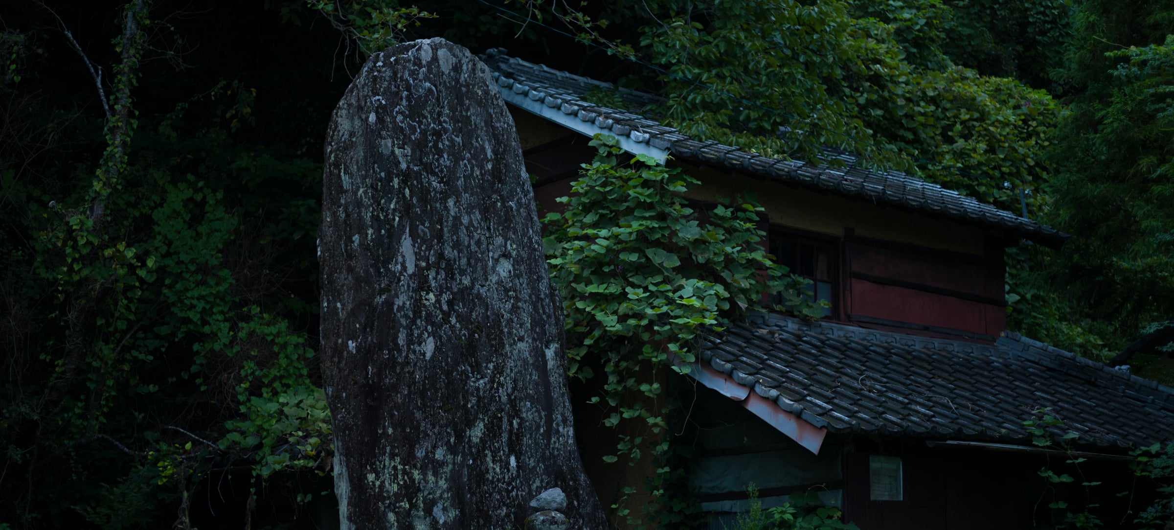 A tall standing stone beside a tile-roofed building, surrounded by greenery (photo: Takahiro Mitsui)