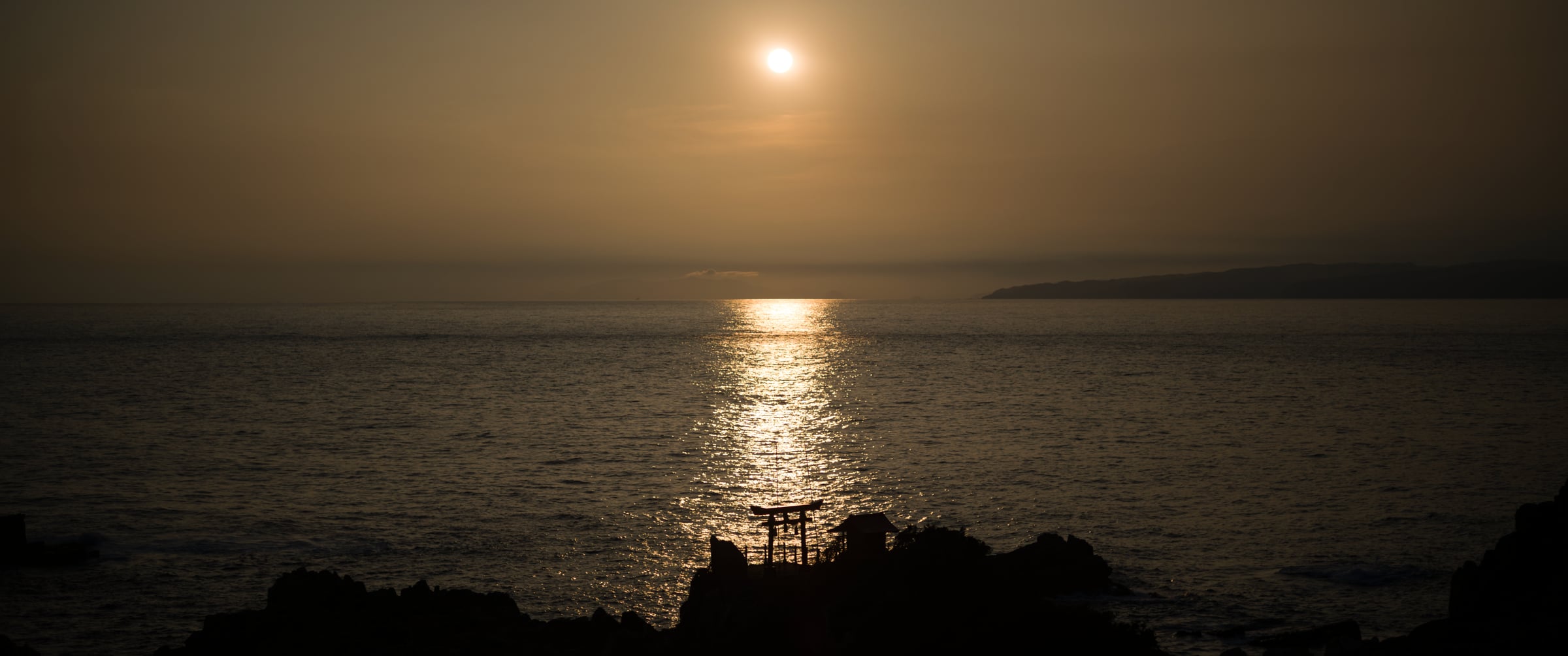 A torii gate silhouetted on the rocks, the sun laying a path of light across the sea (photo: Takahiro Mitsui)