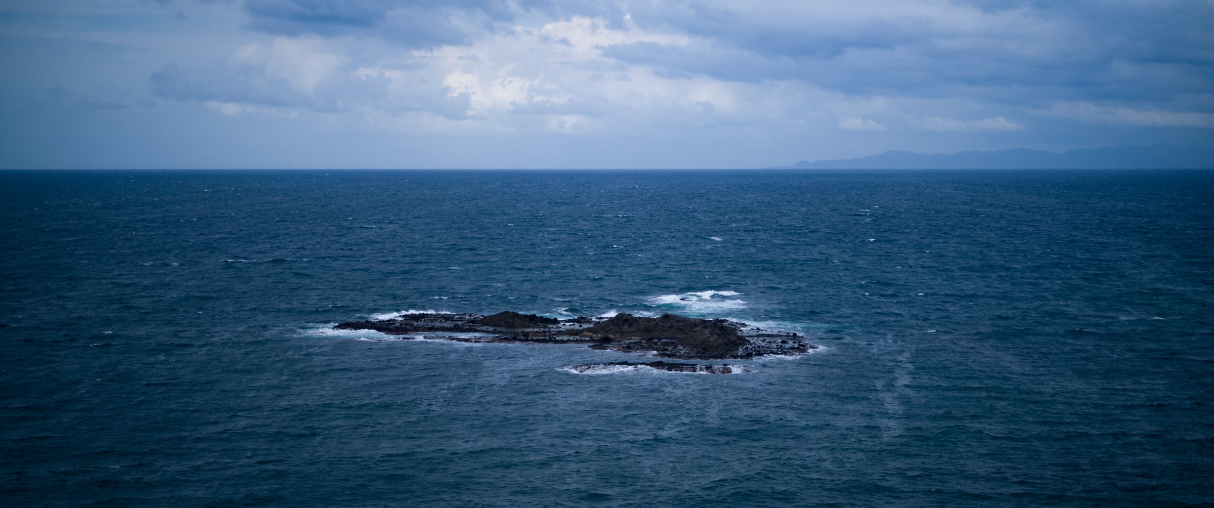 A dark rocky islet in the Sea of Japan, ringed by white surf under a cloudy sky (photo: Takahiro Mitsui)